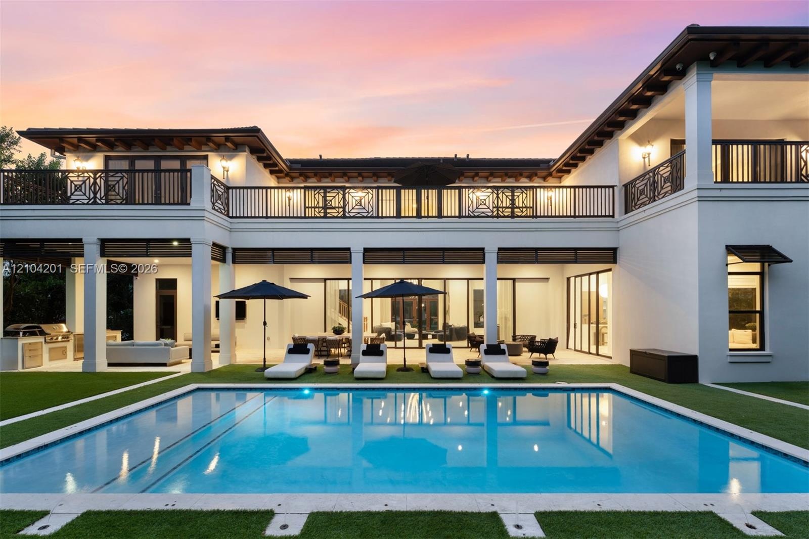 Two-story stucco exterior featuring a rectangular pool, integrated outdoor kitchen, expansive covered terrace, and decorative wrought-iron balcony rai