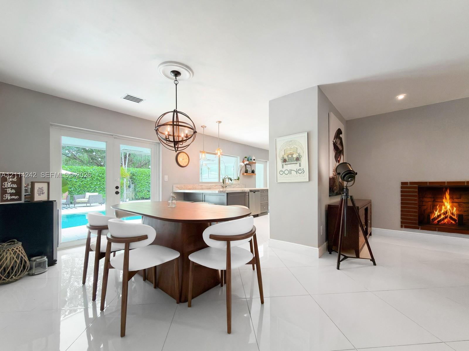 Large-format tile flooring, brick-surround masonry fireplace, spherical metal chandelier, and double-glass doors opening to exterior pool area