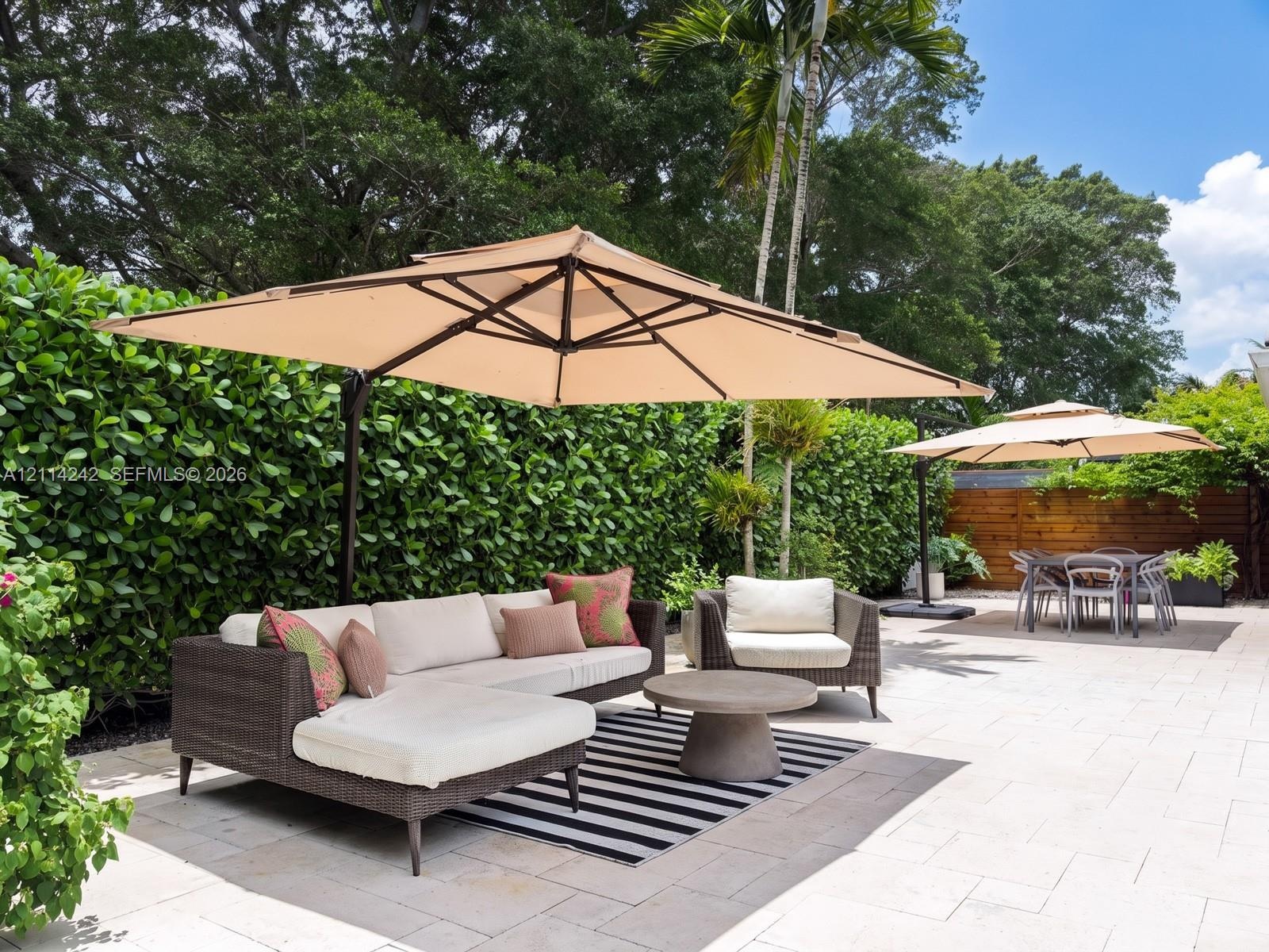 Outdoor patio area featuring light-toned stone pavers, tall perimeter privacy hedging, mature tropical foliage, and natural wood fencing