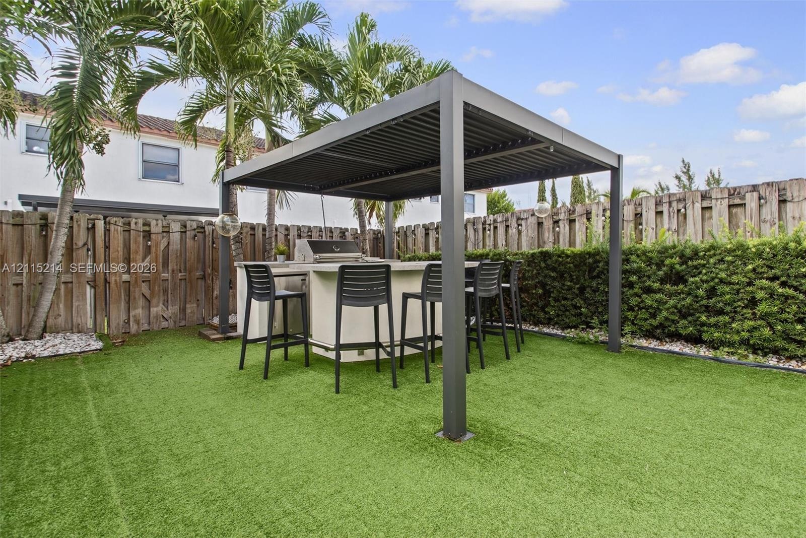 Outdoor living area featuring a covered pergola, integrated built-in grill station with a service counter, and synthetic turf lawn surrounded by matur