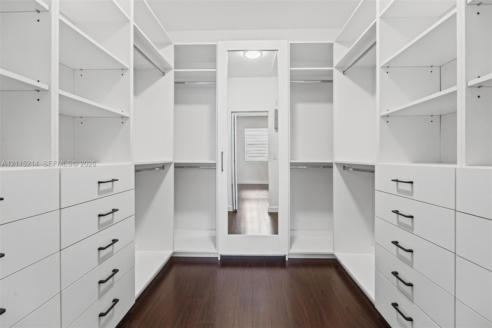 Walk-in closet featuring white built-in cabinetry, floor-to-ceiling shelving, integrated drawers with dark hardware, a center full-length mirror, and 