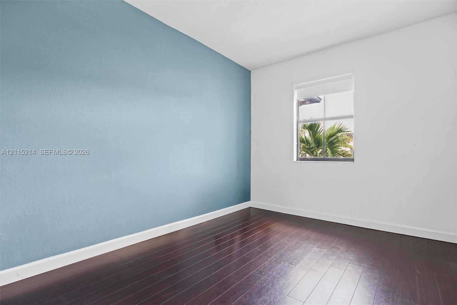 Room featuring wood-finish flooring, a vibrant blue accent wall, and a single-hung window with a roller shade