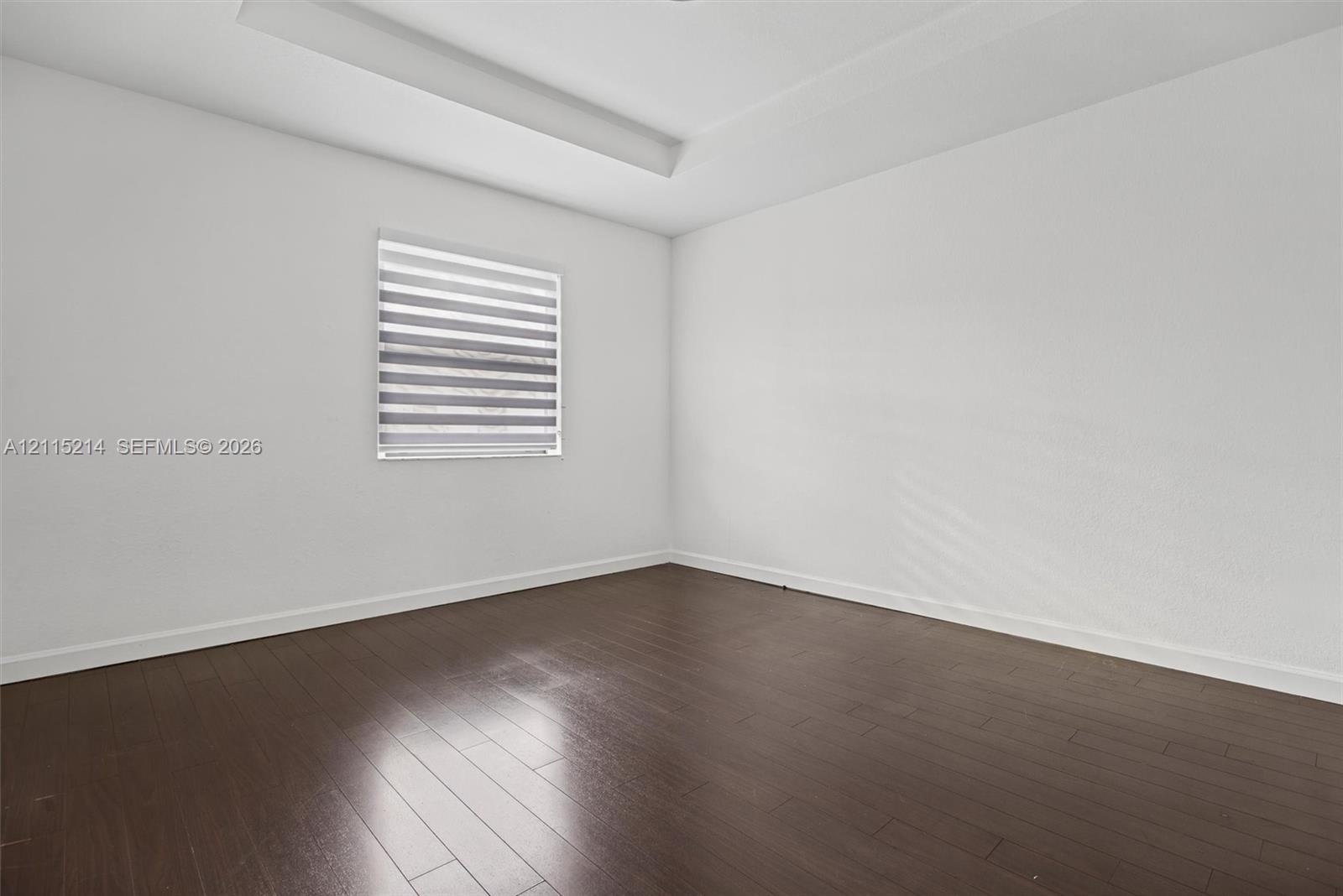 Room featuring dark wood-finish flooring, a tray ceiling, and a window with installed banded roller shades
