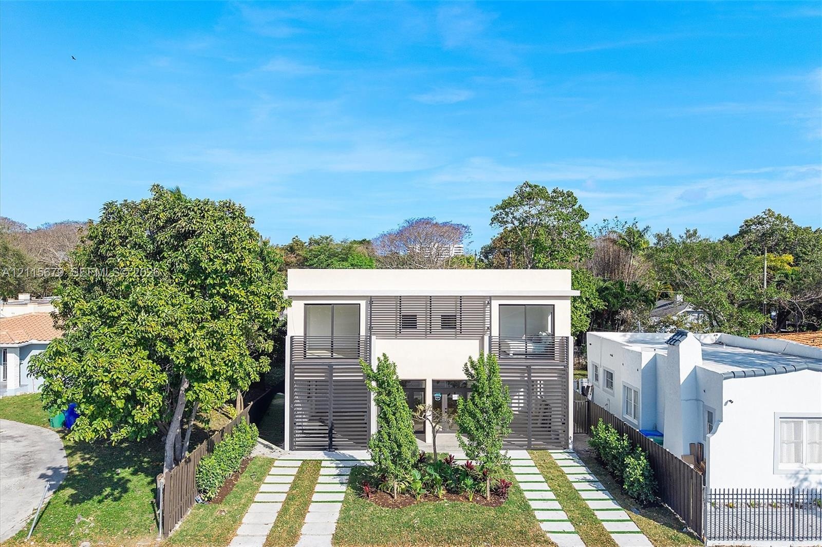 Modern two-story exterior featuring a light-toned stucco facade, horizontal slat privacy screening, double glass balcony doors, and a linear stone-pav