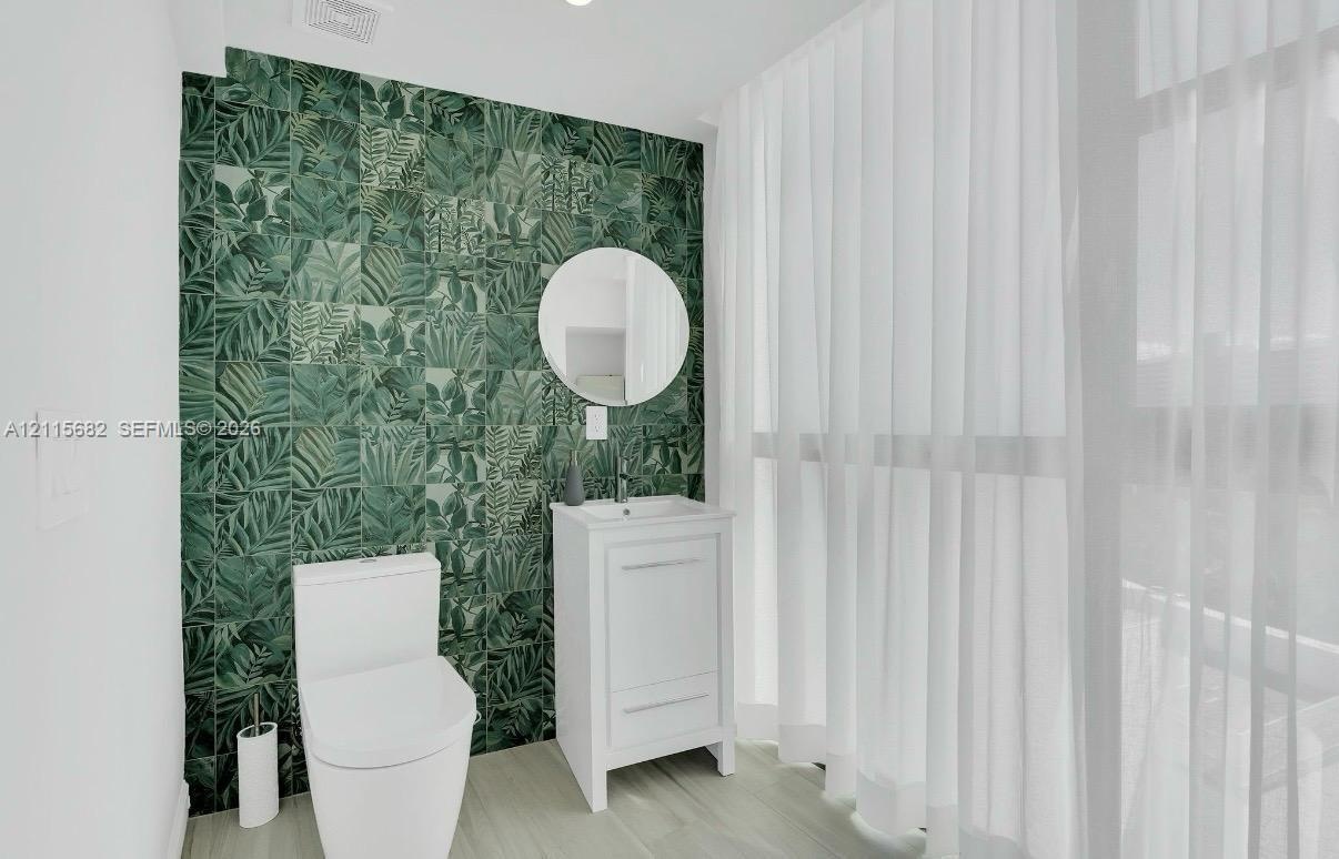 Bathroom featuring a green botanical-patterned tile accent wall, white vanity with an integrated basin, circular wall-mounted mirror, and wood-finish 