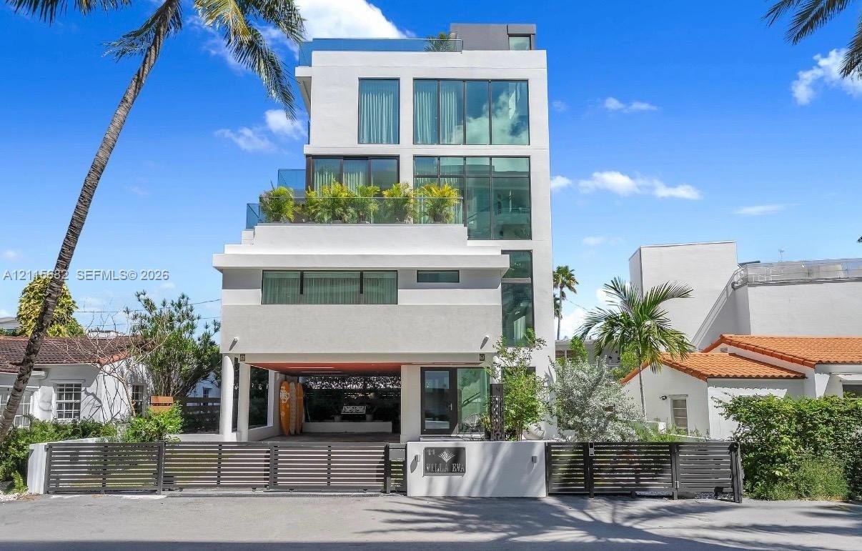 Modern multi-story exterior featuring floor-to-ceiling windows, multiple glass-railed balconies, a gated entrance, and a covered ground-level parking 
