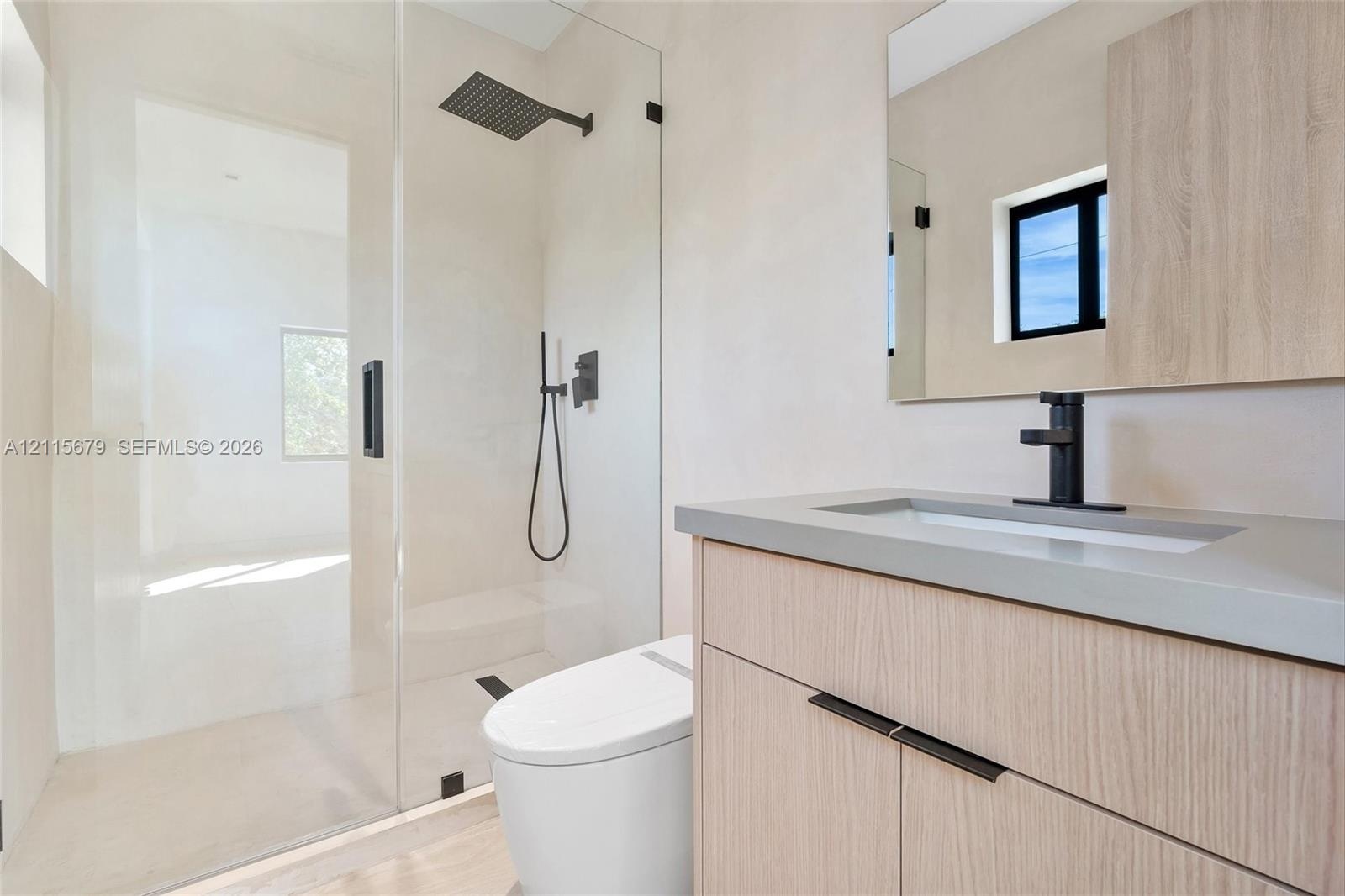 Bathroom featuring a wood-finish vanity with a stone countertop, a glass-enclosed shower with a rainfall showerhead, a wall-mounted mirror, and matte 