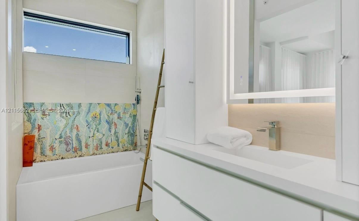Floating white vanity with solid-surface countertop, integrated LED-backlit mirror, soaking tub featuring a decorative mosaic tile surround, and horiz