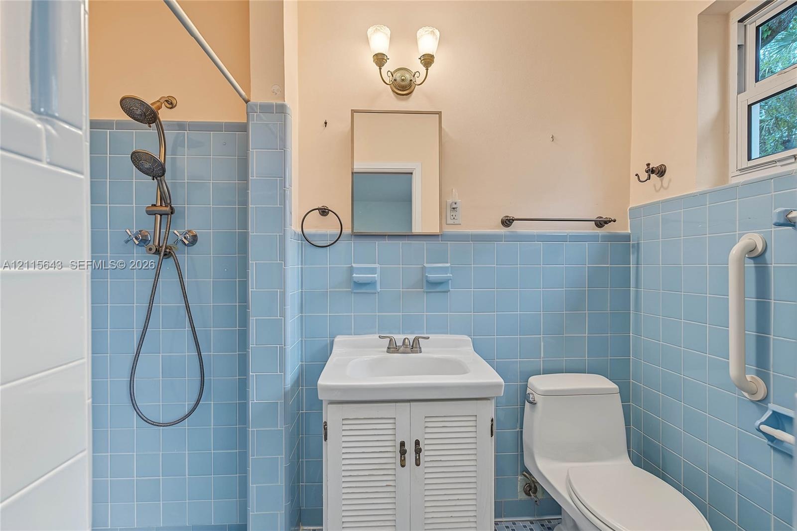 Bathroom featuring blue tile wainscoting, pedestal-style vanity with louvered cabinet doors, dual-head shower fixture, wall-mounted light fixture, and