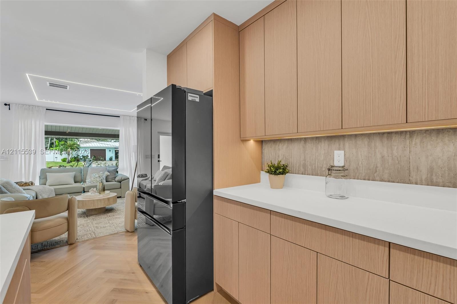 Flat-panel light wood cabinetry featuring under-cabinet lighting, solid surface countertops, textured backsplash, and herringbone-pattern wood-finish 