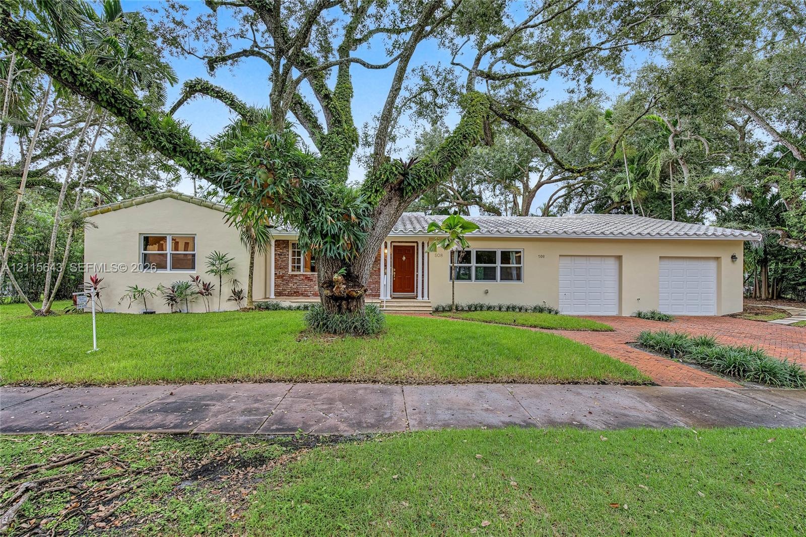 Single-story residence featuring a tile roof, brick masonry facade, two-car garage, and mature canopy trees on a landscaped lot