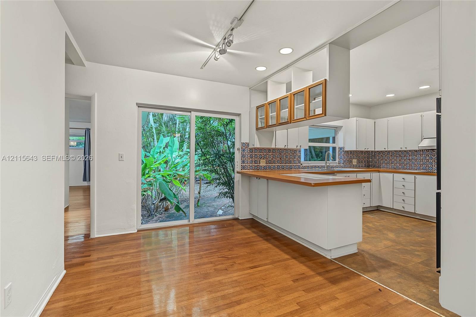 Kitchen featuring wood-finish flooring, white cabinetry with glass-front uppers, wood-tone countertops, patterned tile backsplash, and sliding glass d