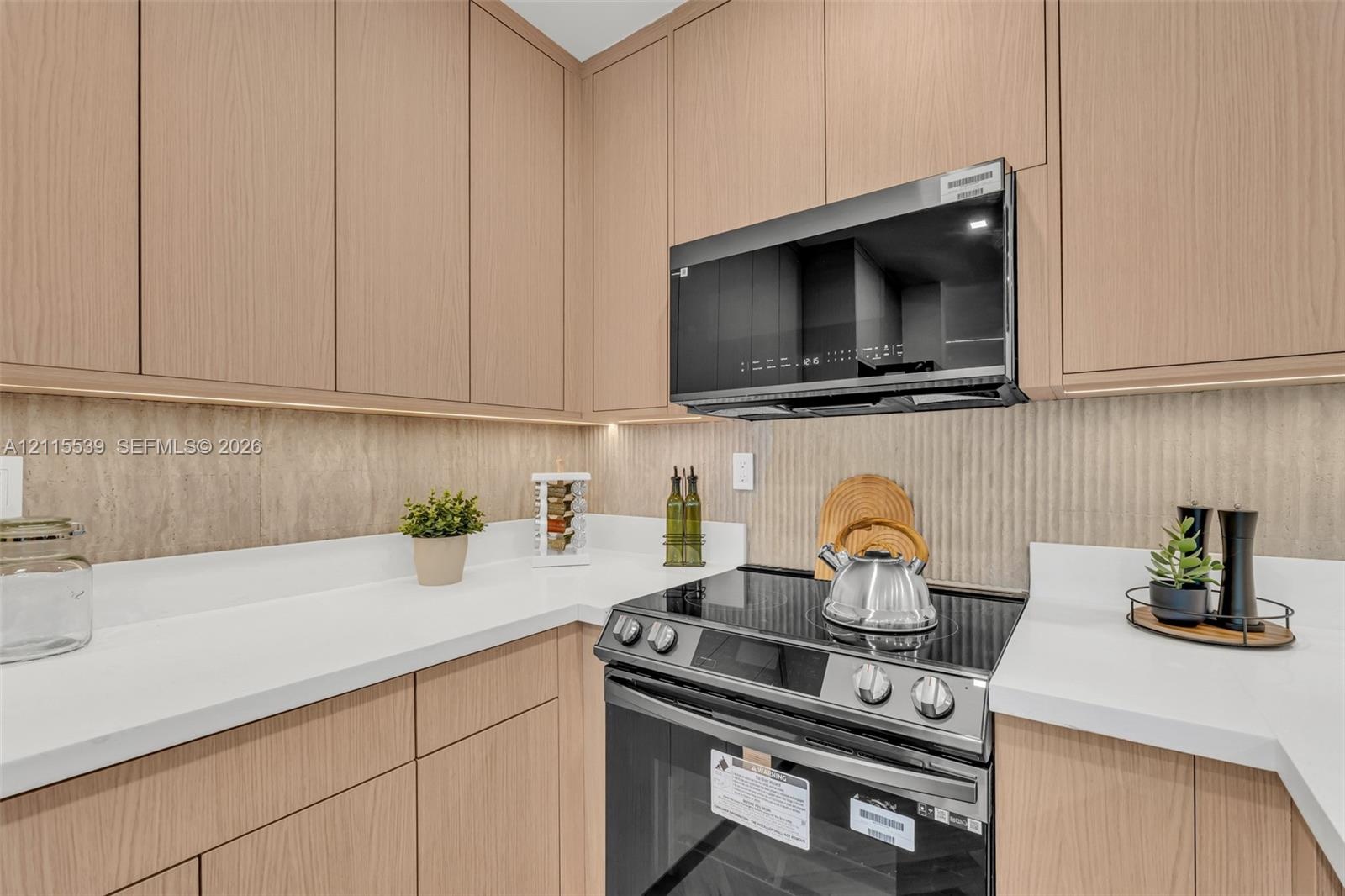 Flat-panel light wood cabinetry featuring under-cabinet lighting, solid surface countertops, a textured tile backsplash, and stainless steel integrate