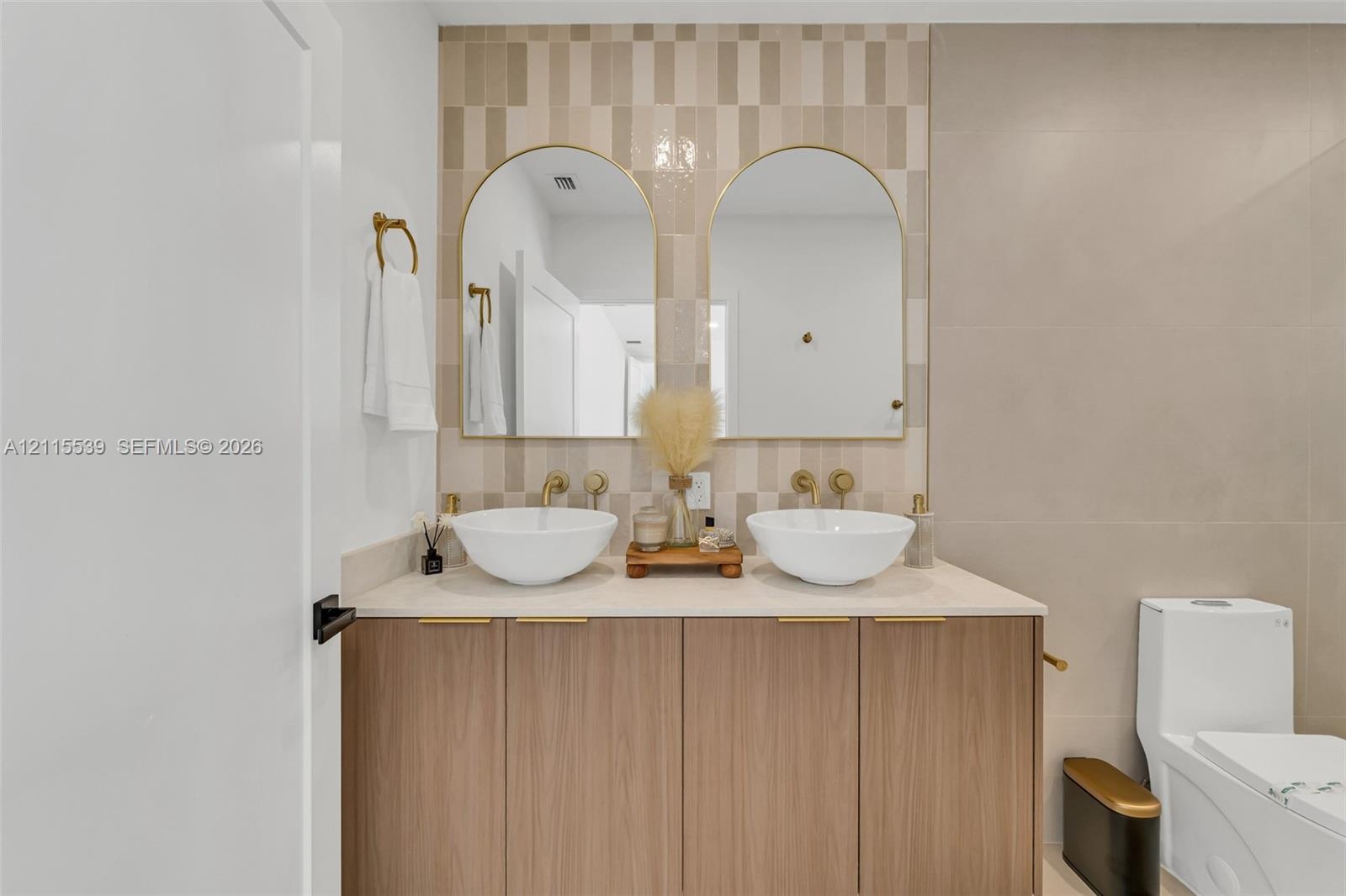 Double vanity featuring wood-finish cabinetry, white vessel sinks, gold-tone wall-mounted faucets, arched mirrors, and a patterned tile backsplash