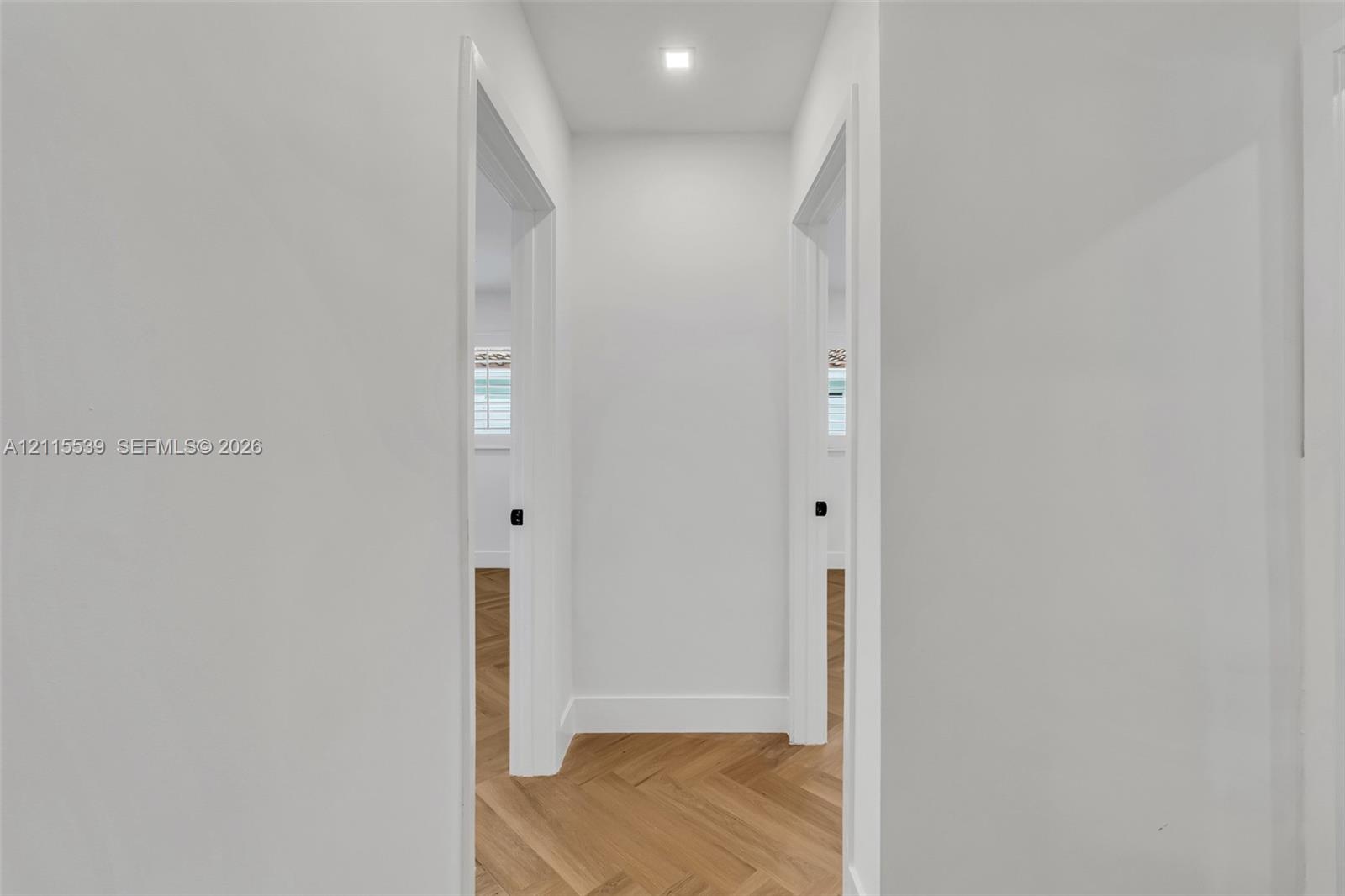 Interior hallway featuring light wood-finish flooring in a herringbone pattern, smooth white walls, recessed ceiling lighting, and matte black door ha