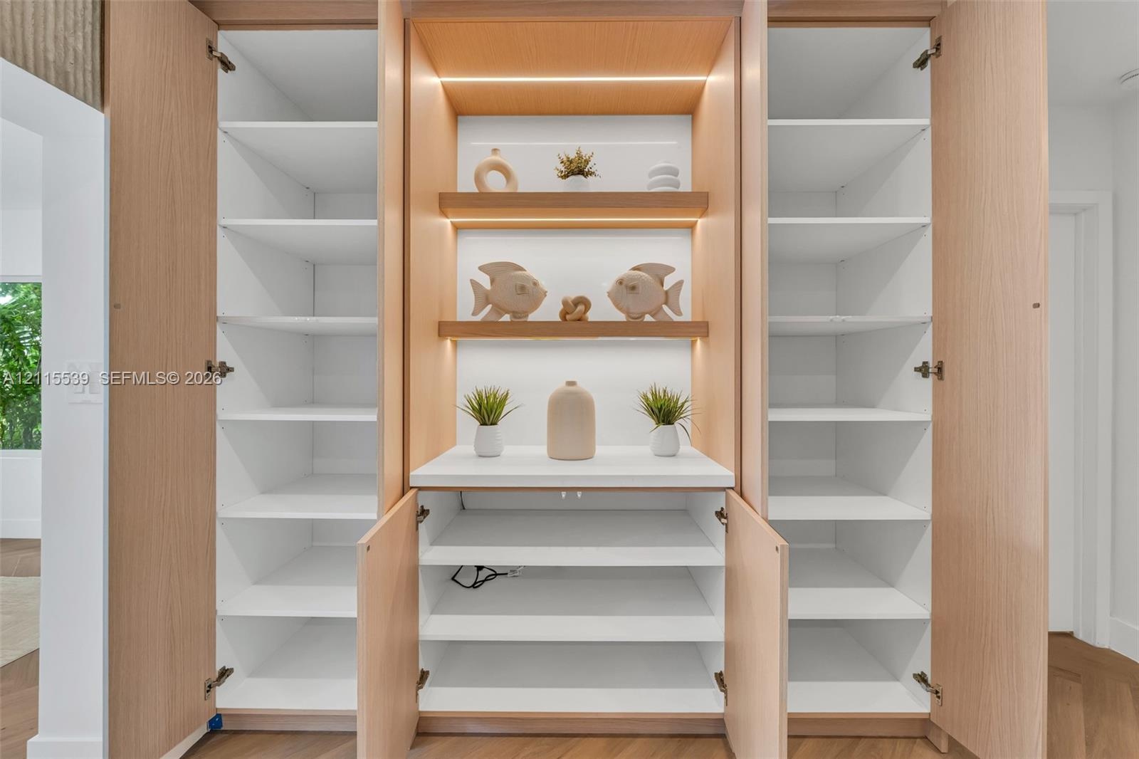 Custom built-in cabinetry featuring light wood-finish veneer, integrated LED shelf lighting, fixed white shelving, and a central solid-surface counter