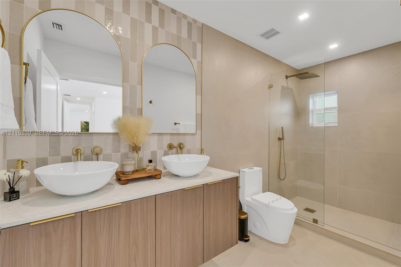 Dual vanity featuring vessel sinks, quartz countertops, wood-finish cabinetry, arched mirrors with gold-toned frames, and a walk-in shower with a glas