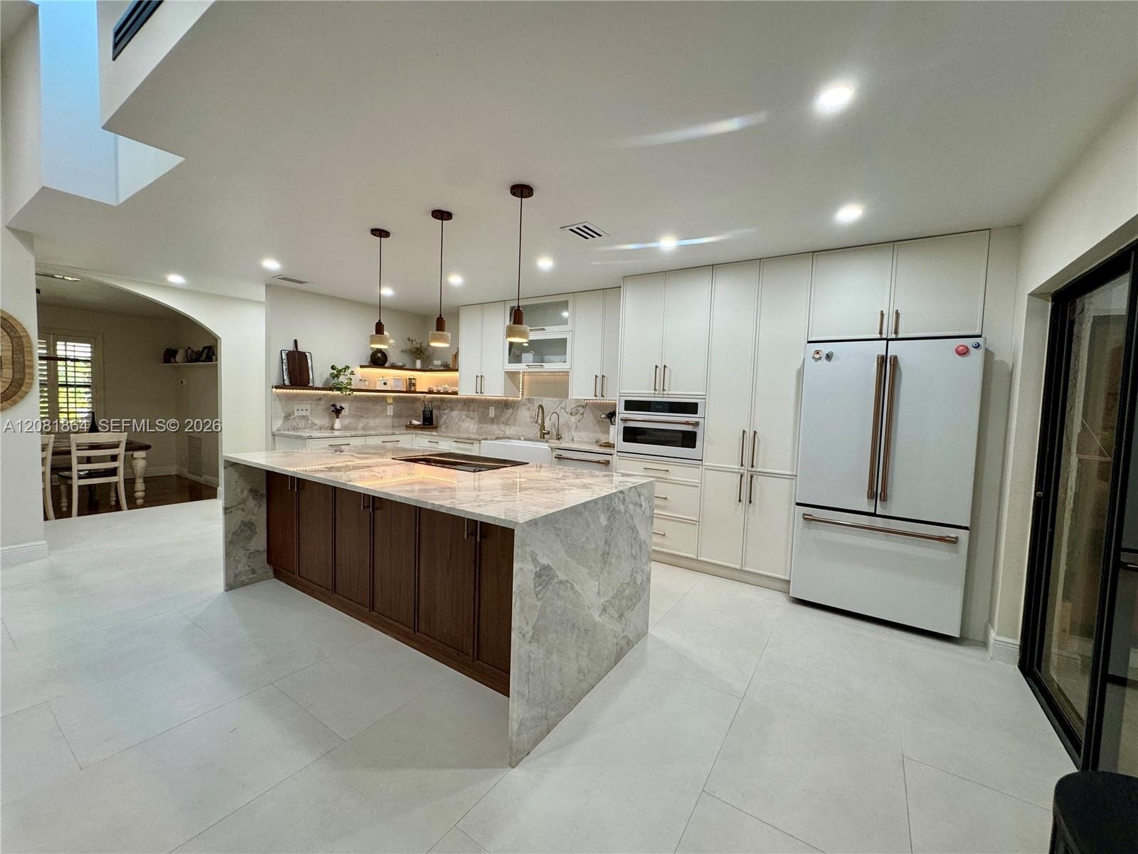 Kitchen featuring a large central island with a waterfall countertop, white cabinetry, a farmhouse-style apron sink, integrated lighting, and three pe