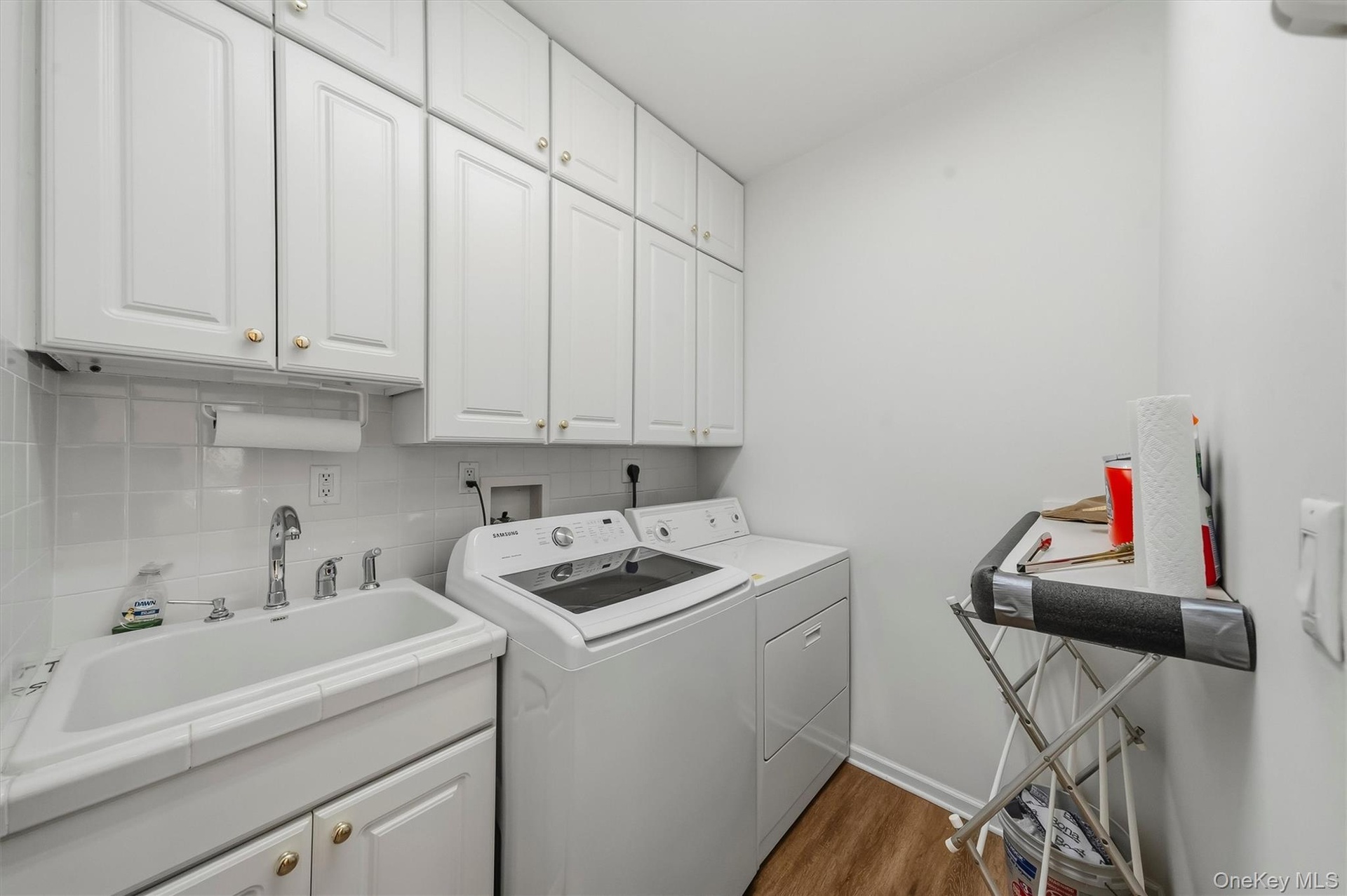 White cabinetry with gold-tone hardware, utility sink with chrome  faucet, white square tile backsplash, wood-finish flooring