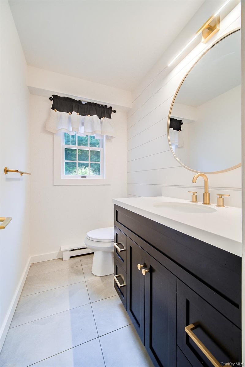 First floor 1/2 bath features wood-finish vanity with gold-tone hardware, white stone countertop, round gold-tone framed mirror, tiled flooring and wh