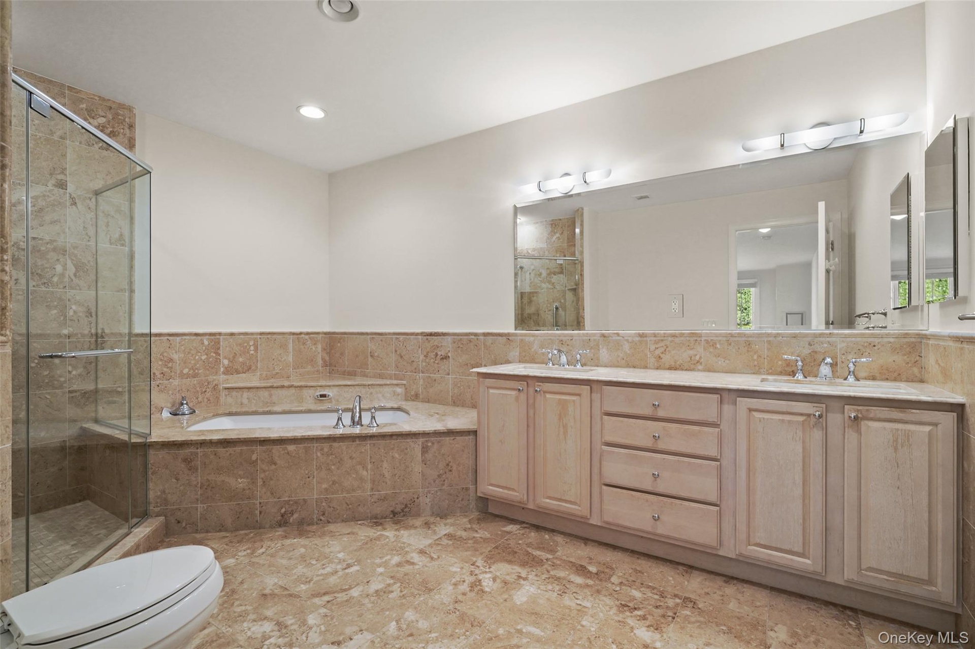 Travertine tile flooring, built-in bathtub, glass-enclosed shower, dual vanity with undermount sinks, and a large wall-mounted mirror