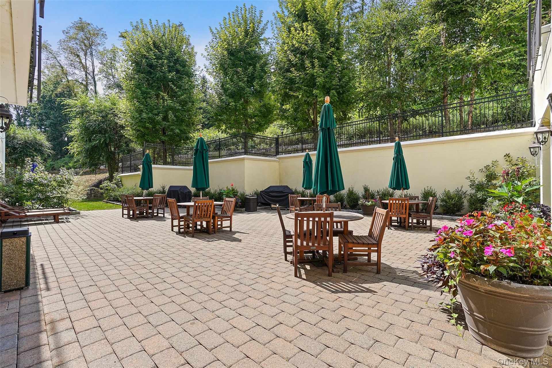 Expansive paver patio with a low retaining wall, black wrought iron fencing, mature trees, and landscaping