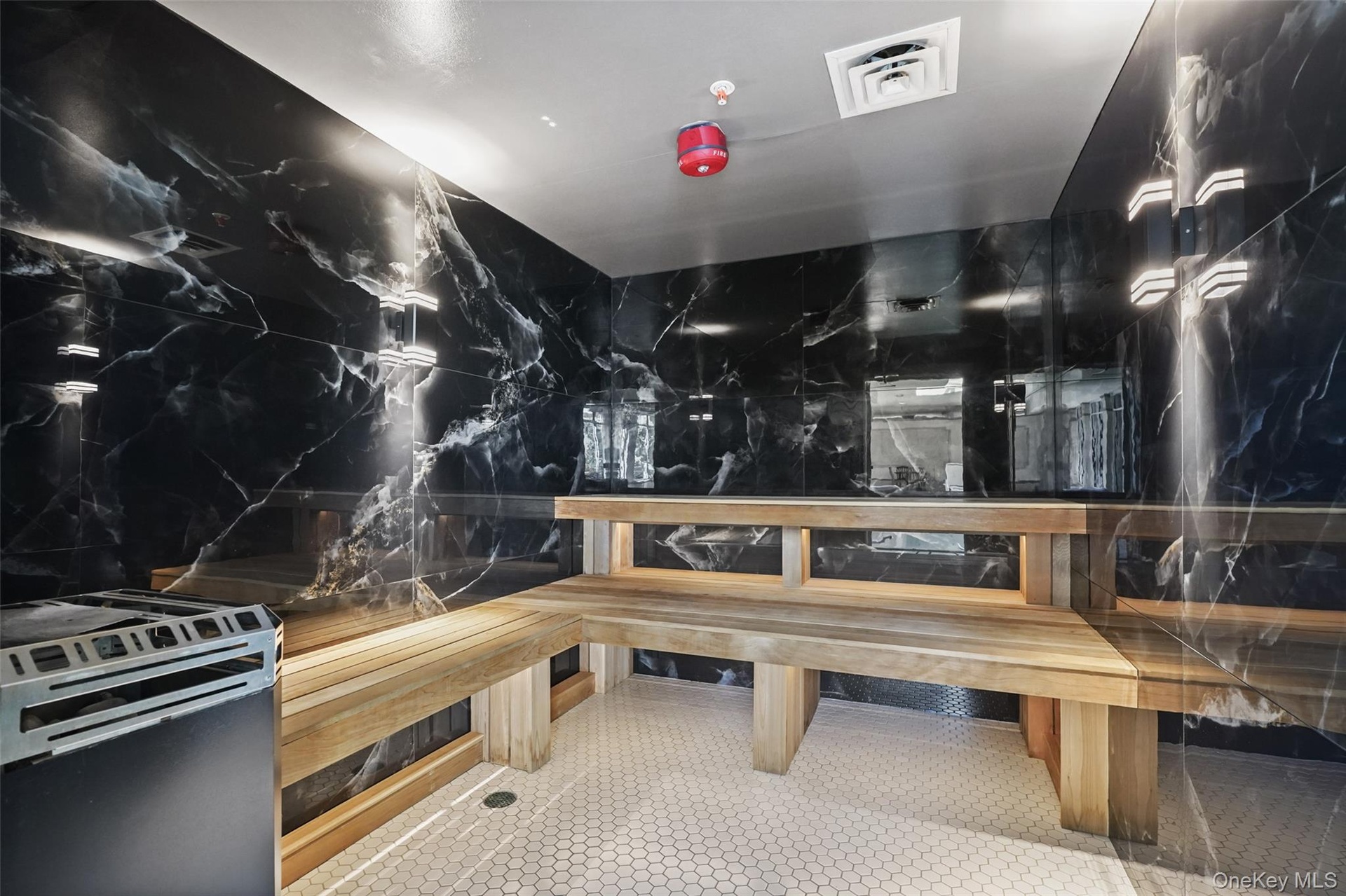 Black marble-style wall finishes, light wood benches, white hexagonal floor tiling, and a built-in heating unit