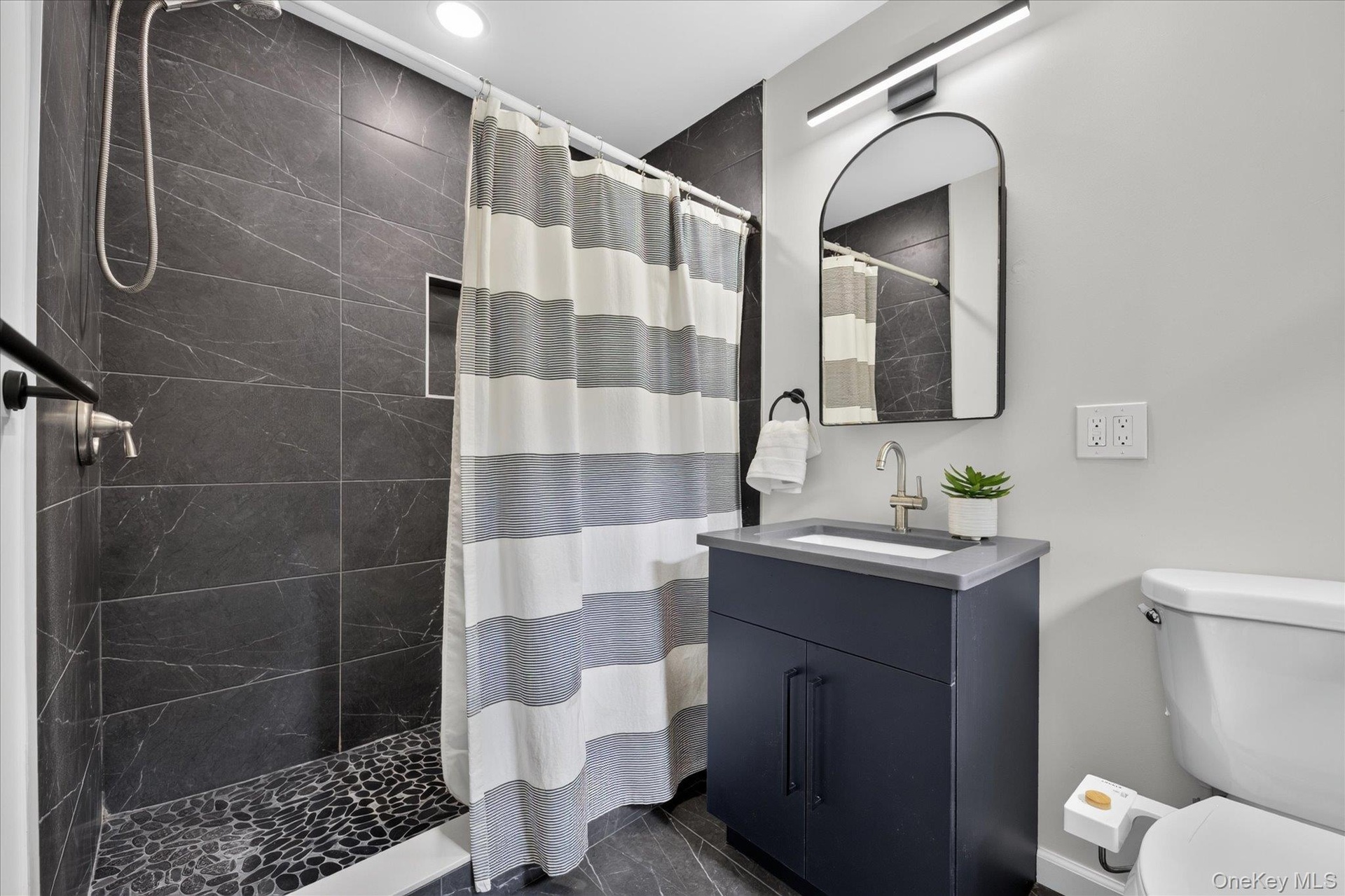 Shower with dark tile surround and pebble floor, dark vanity cabinet with light countertop, arched mirror, and contemporary light fixture