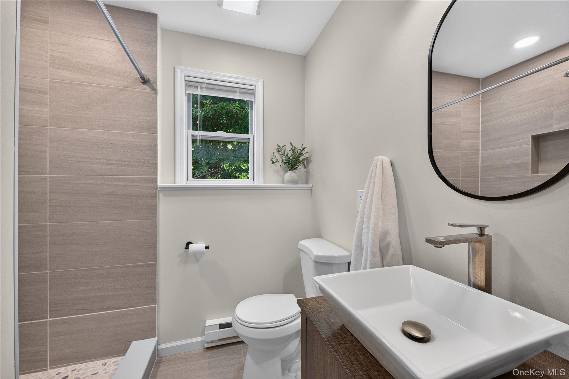 Lower-level full bathroom, Modern bathroom featuring a rectangular vessel sink, wood-finish vanity, large oval mirror, tiled shower with recessed shel