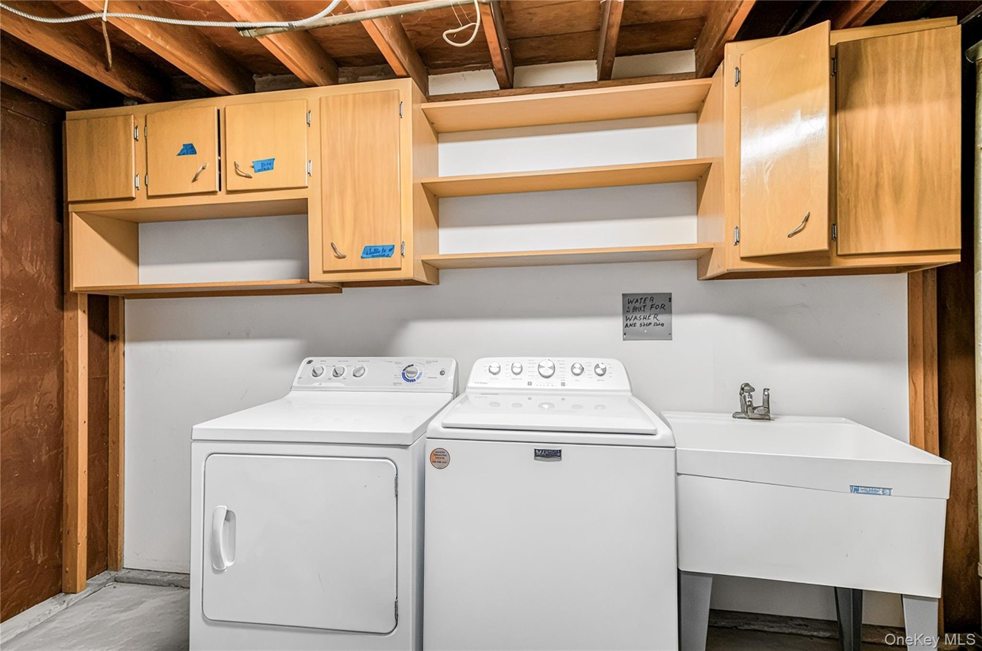 Utility room with exposed ceiling joists, light wood-finish cabinetry, open shelving, and a utility sink
