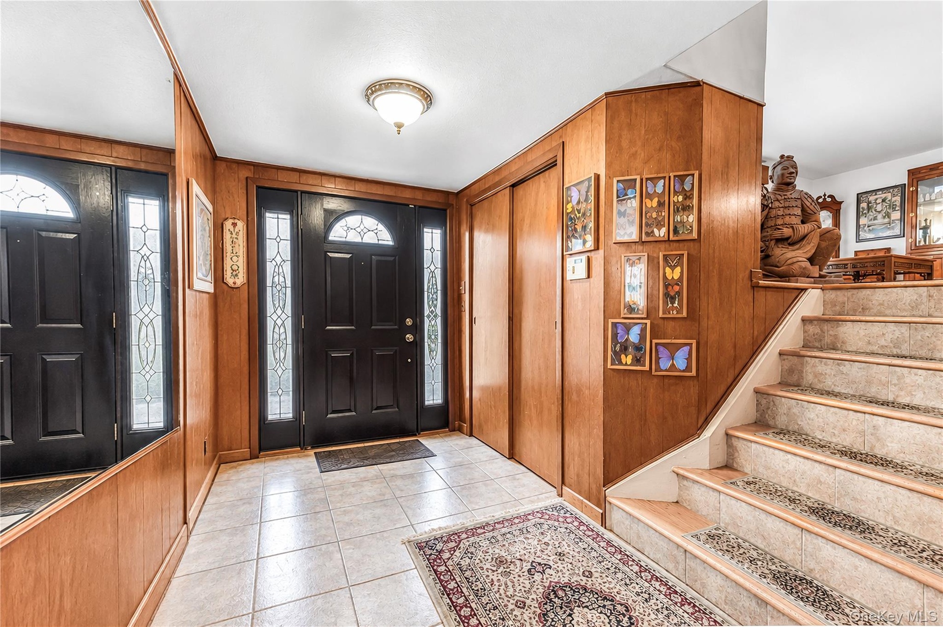 Foyer featuring a black entry door with decorative glass sidelights, tile flooring, wood paneling, and a staircase with an integrated runner