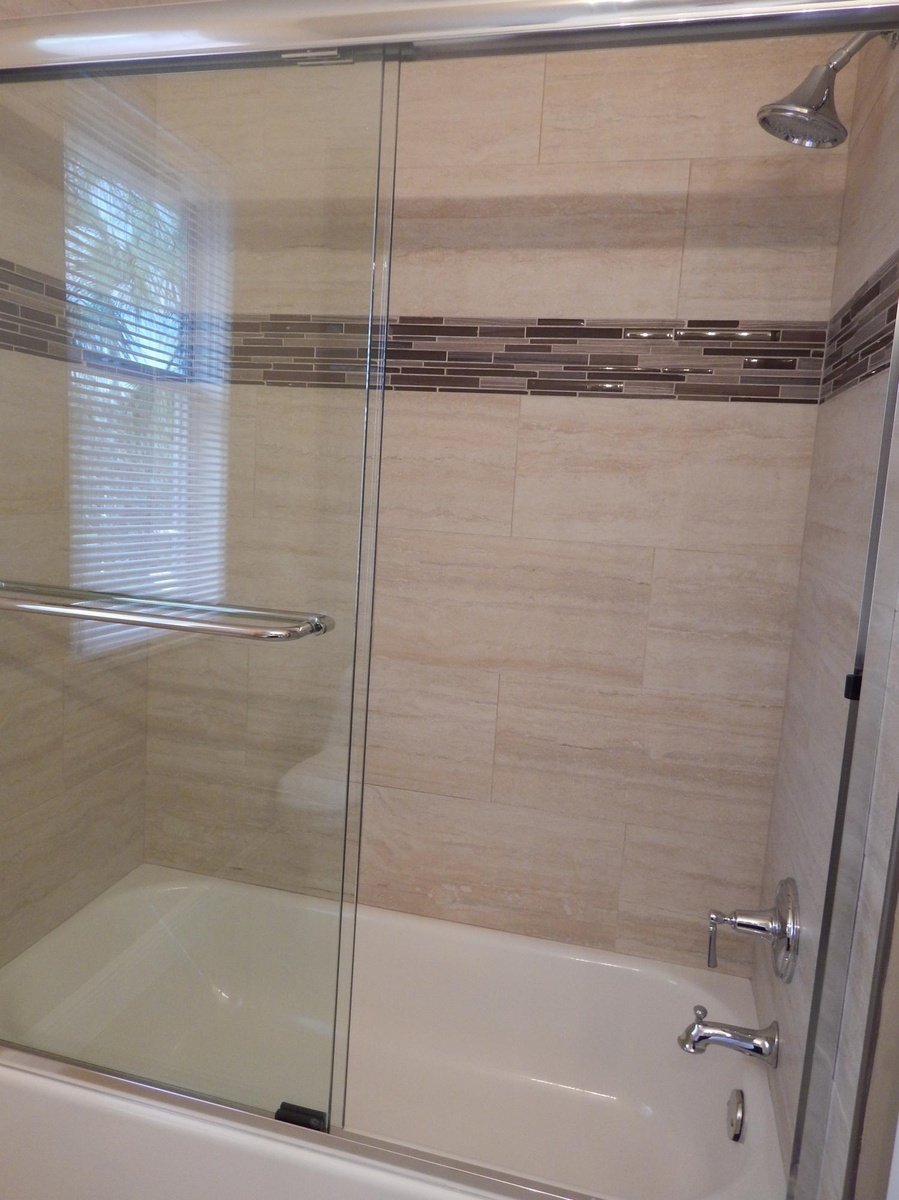 Bathtub with chrome fixtures, full-height tile surround, decorative tile accent band, and clear glass sliding shower doors