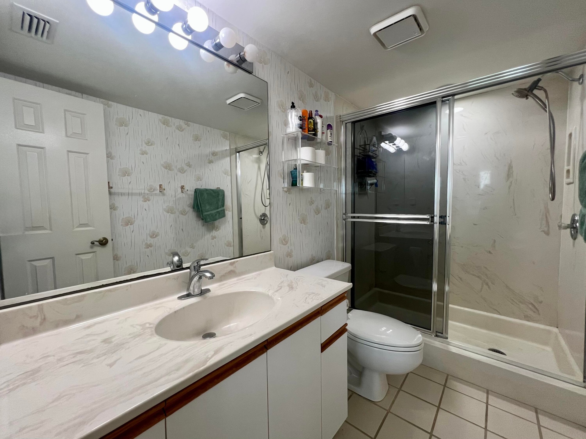 Vanity with integrated sink and chrome faucet, full-width mirror with vanity lighting, white paneled cabinetry, tiled flooring, and a glass-enclosed s