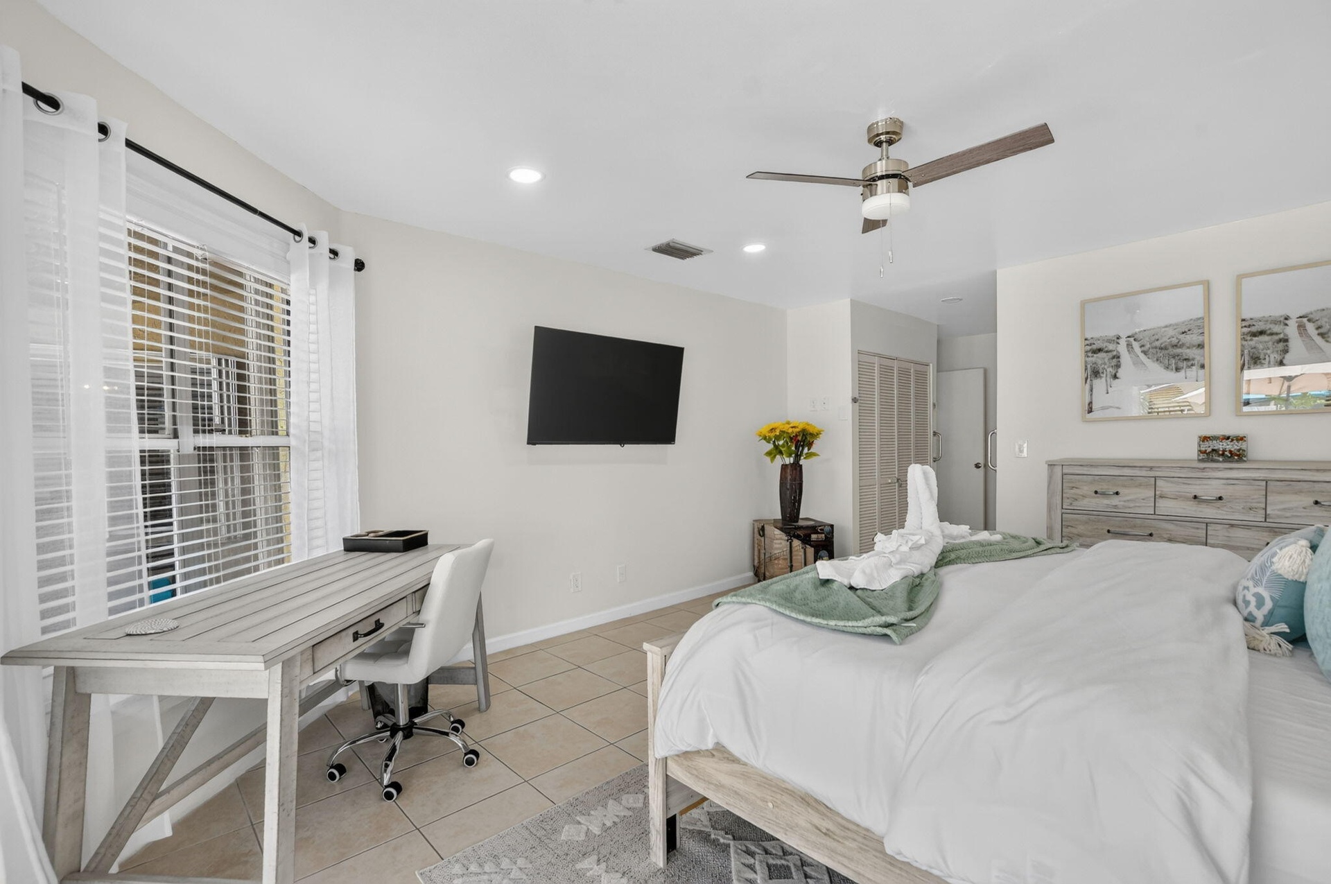 Spacious primary bedroom featuring tile flooring, a ceiling fan with integrated lighting, a wall-mounted flat-screen television, and a dedicated work 