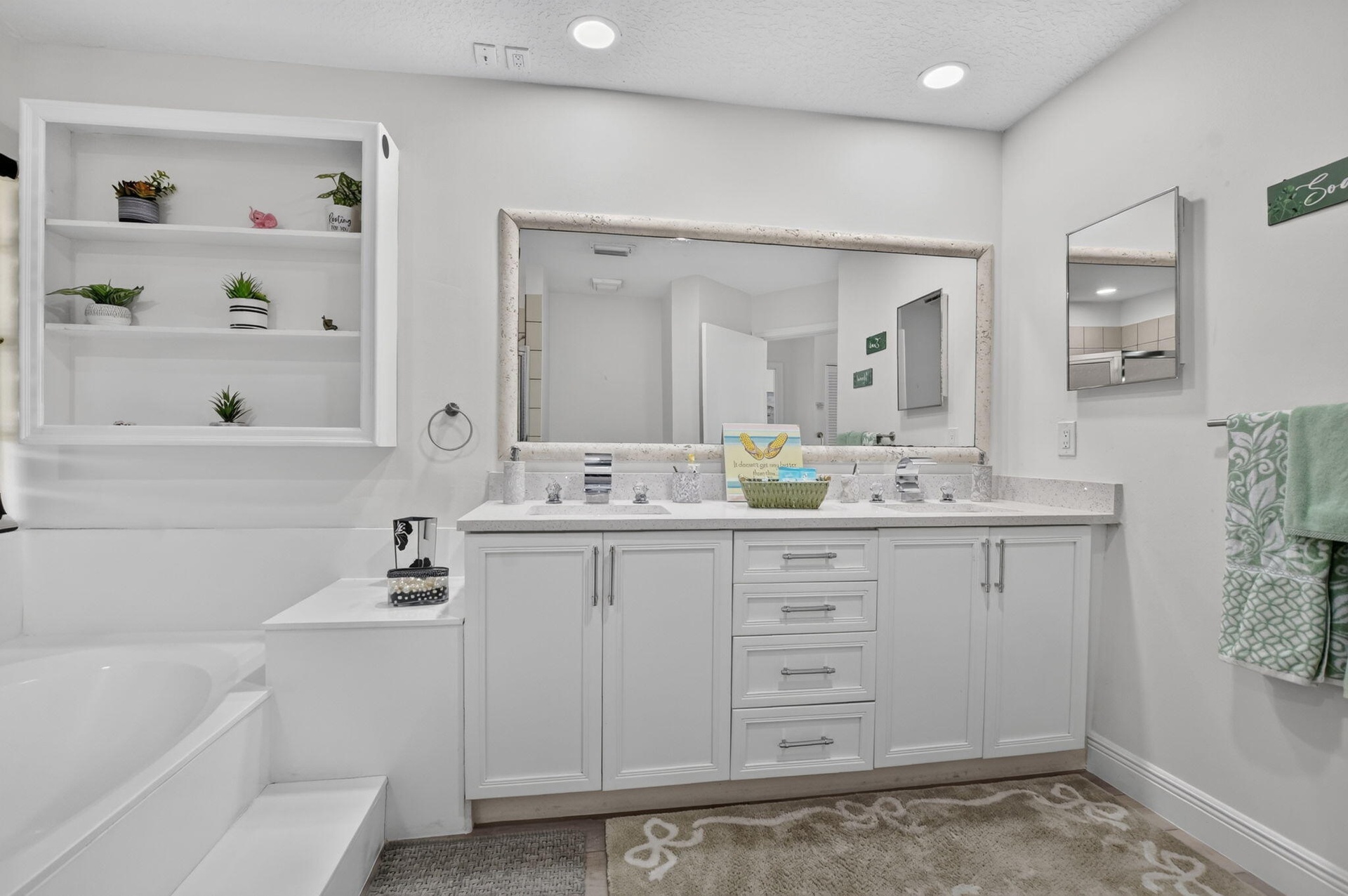 Dual basin vanity with white cabinetry and stone-finish countertop, large framed mirror, recessed lighting, built-in shelving, and a large soaking tub