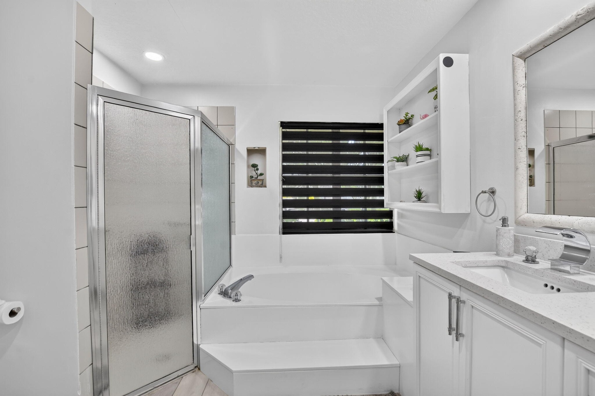 Built-in bathtub with integrated steps, frosted glass shower enclosure, white vanity with integrated sink, stone-finish countertop, and wall-mounted s