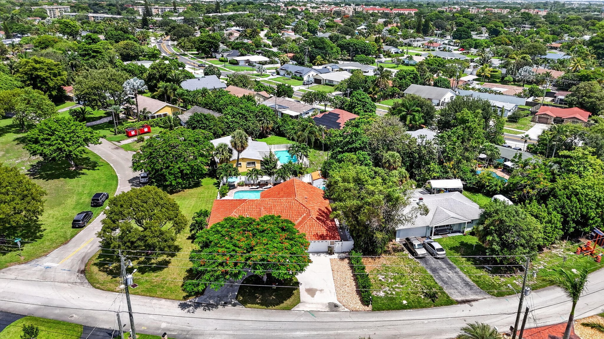 Aerial view showcasing a substantial corner-lot property with a clay tile roof, mature landscaping throughout the grounds, a sparkling swimming pool w