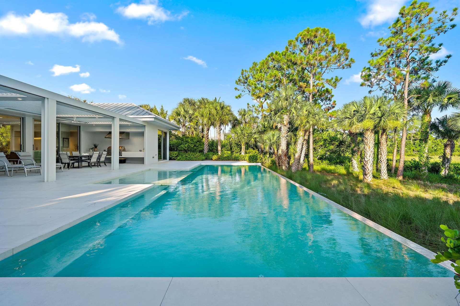 Expansive outdoor area featuring an infinity-edge pool with light-colored coping, a covered patio with a built-in outdoor kitchen, and tropical landsc