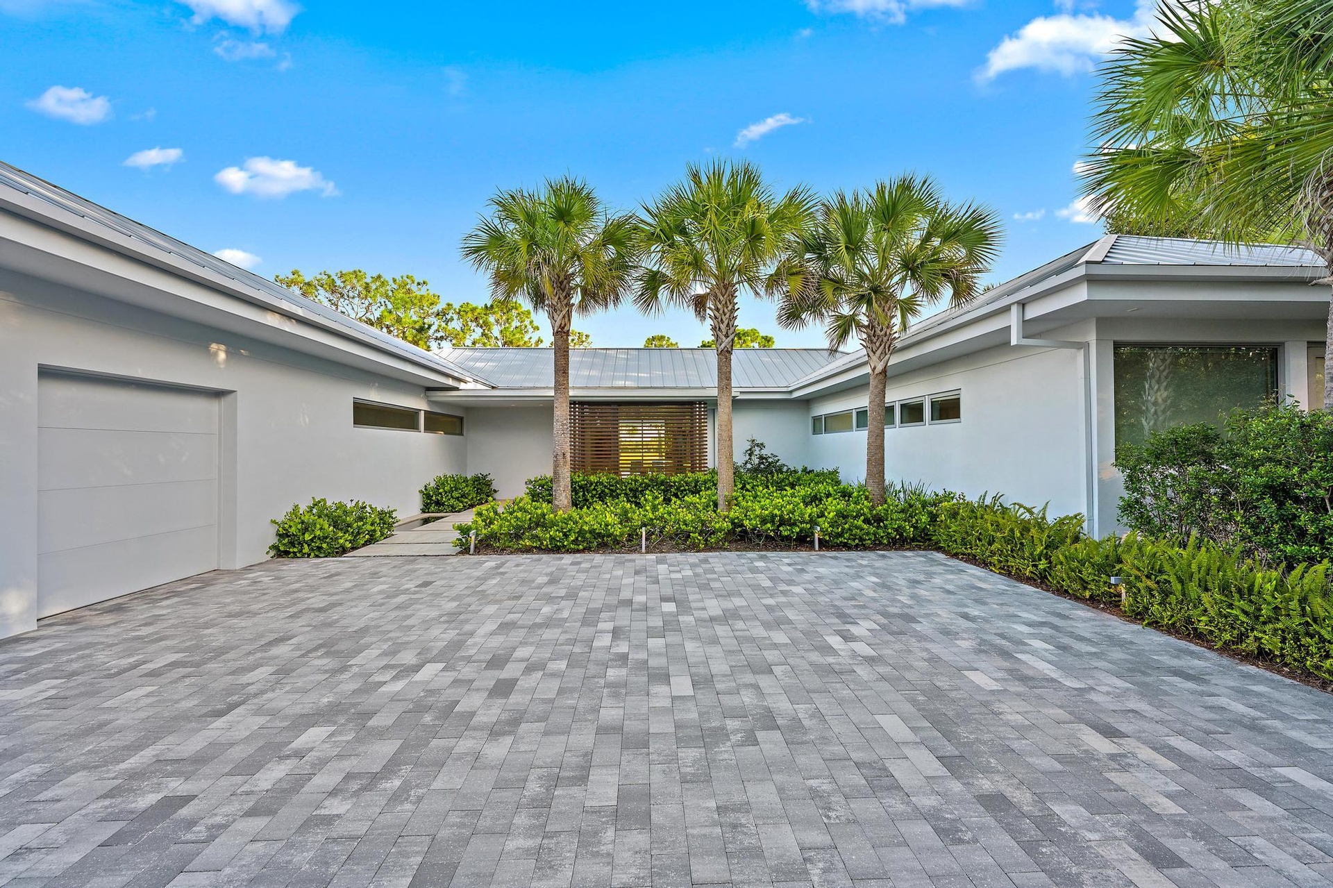 Elegant driveway featuring permeable pavers in a geometric pattern, bordered by lush tropical landscaping with mature palm trees and manicured hedges