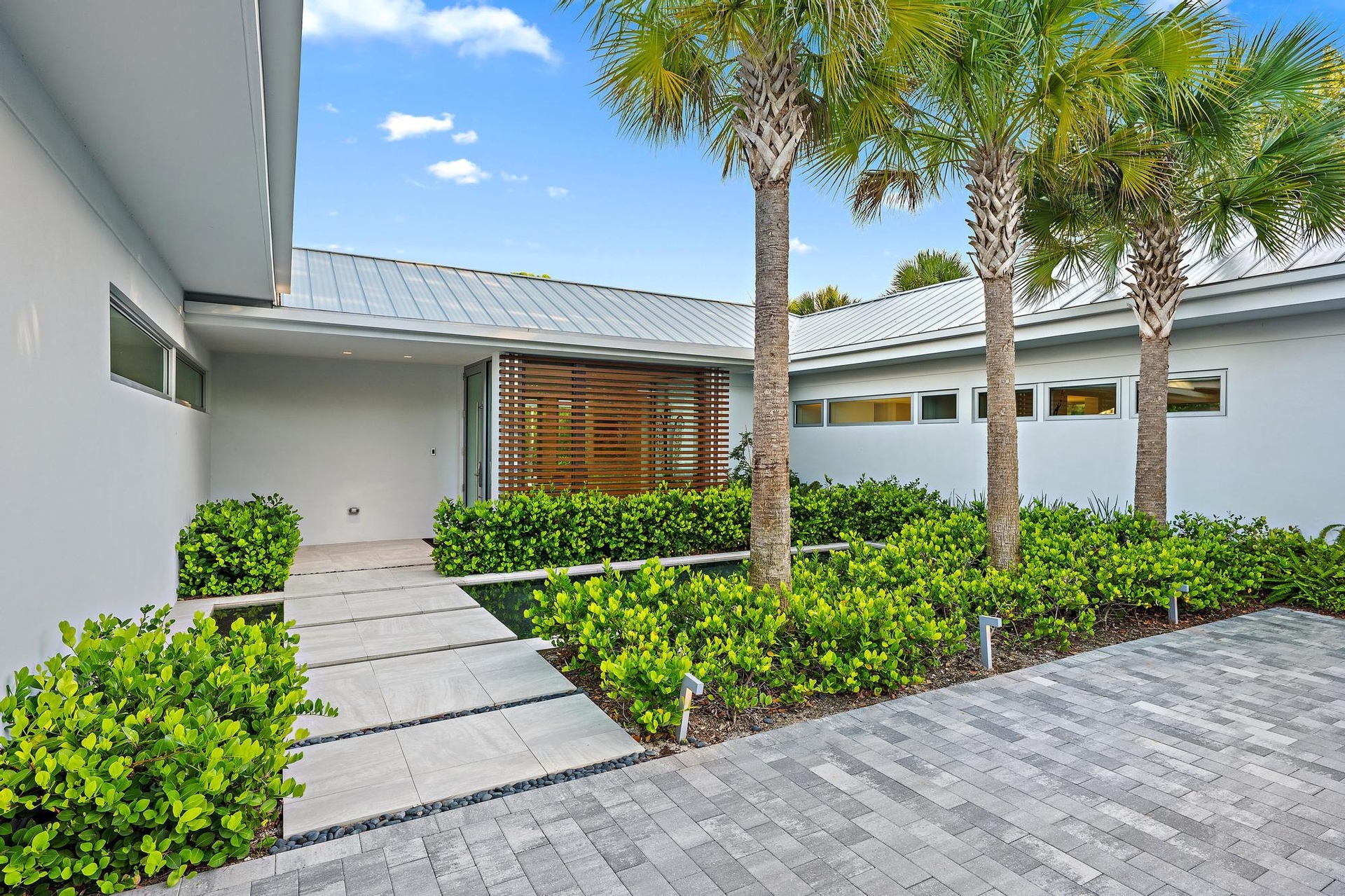 Paver driveway, mature palm trees, contemporary exterior, metal roof, and lush landscaping