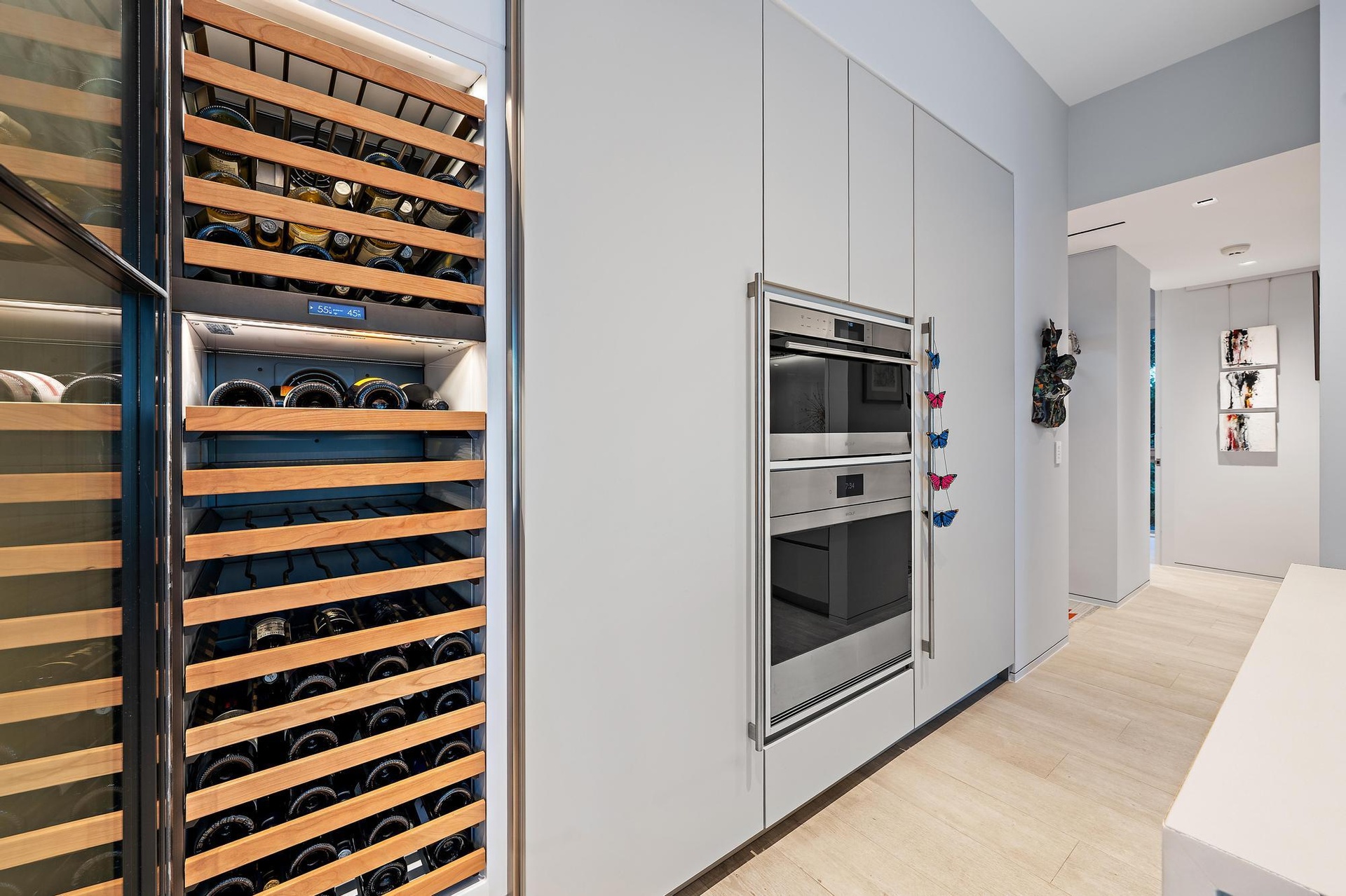 Integrated wine refrigerator with wood racks, built-in double ovens with stainless steel finishes, light wood-finish flooring, and minimalist flat-pan