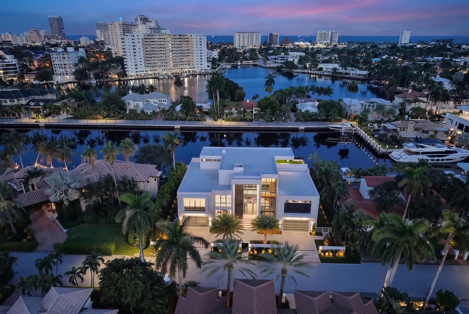 A breathtaking twilight aerial showcases the residence’s illuminated modern architecture, tropical landscaping, and exceptional waterfront setting, fr