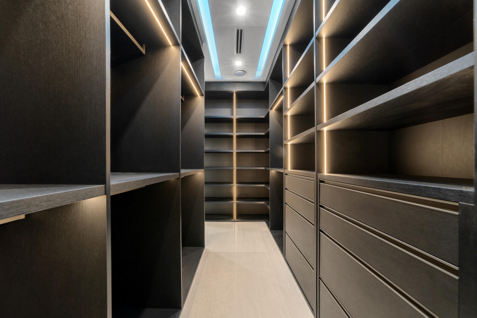 Custom gentleman’s walk-in wardrobe featuring dark wood cabinetry, extensive shelving and hanging space, integrated drawers, and sophisticated lightin