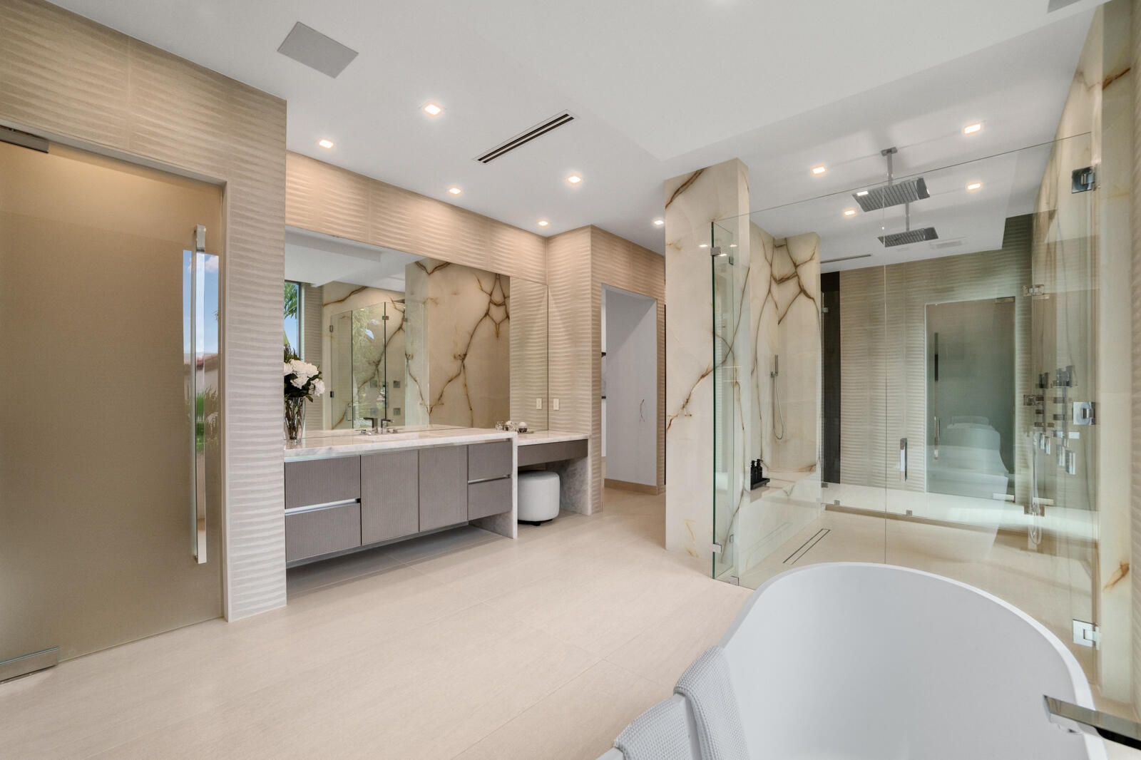 Luxurious primary bath showcasing an oversized glass-enclosed shower with dual rain heads, book-matched stone, floating vanities, and a dedicated make