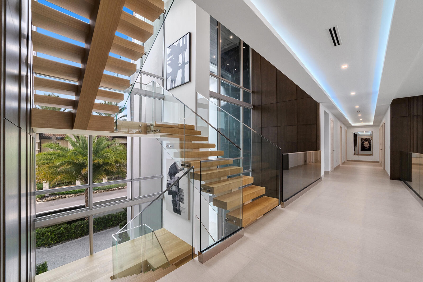 Second-floor landing showcasing the floating staircase from both sides, framed by soaring windows, glass railings, and rich wood-paneled accent walls.