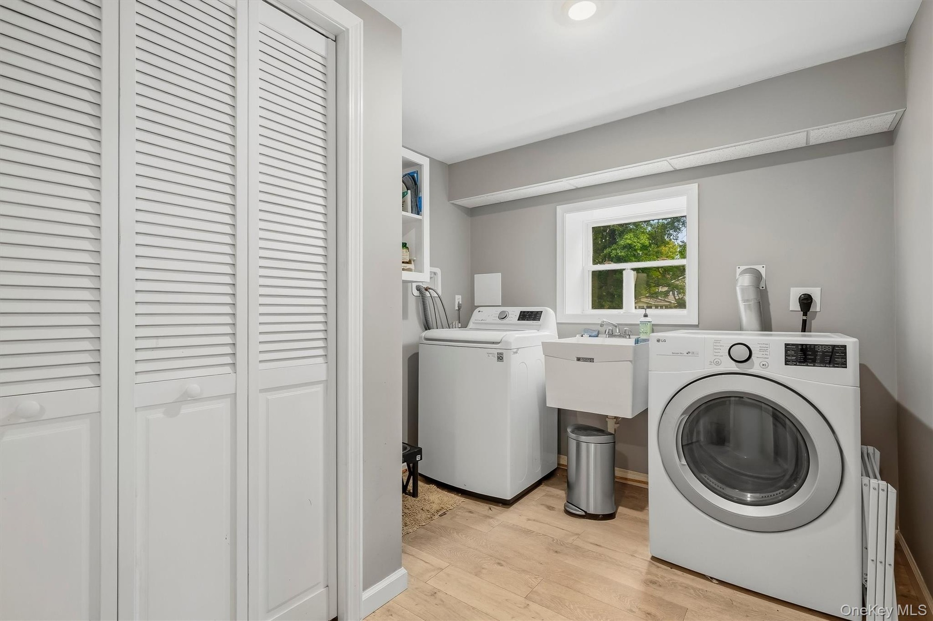 White louvered bifold doors, window with natural light, utility sink, overhead lighting, and wood-finish flooring