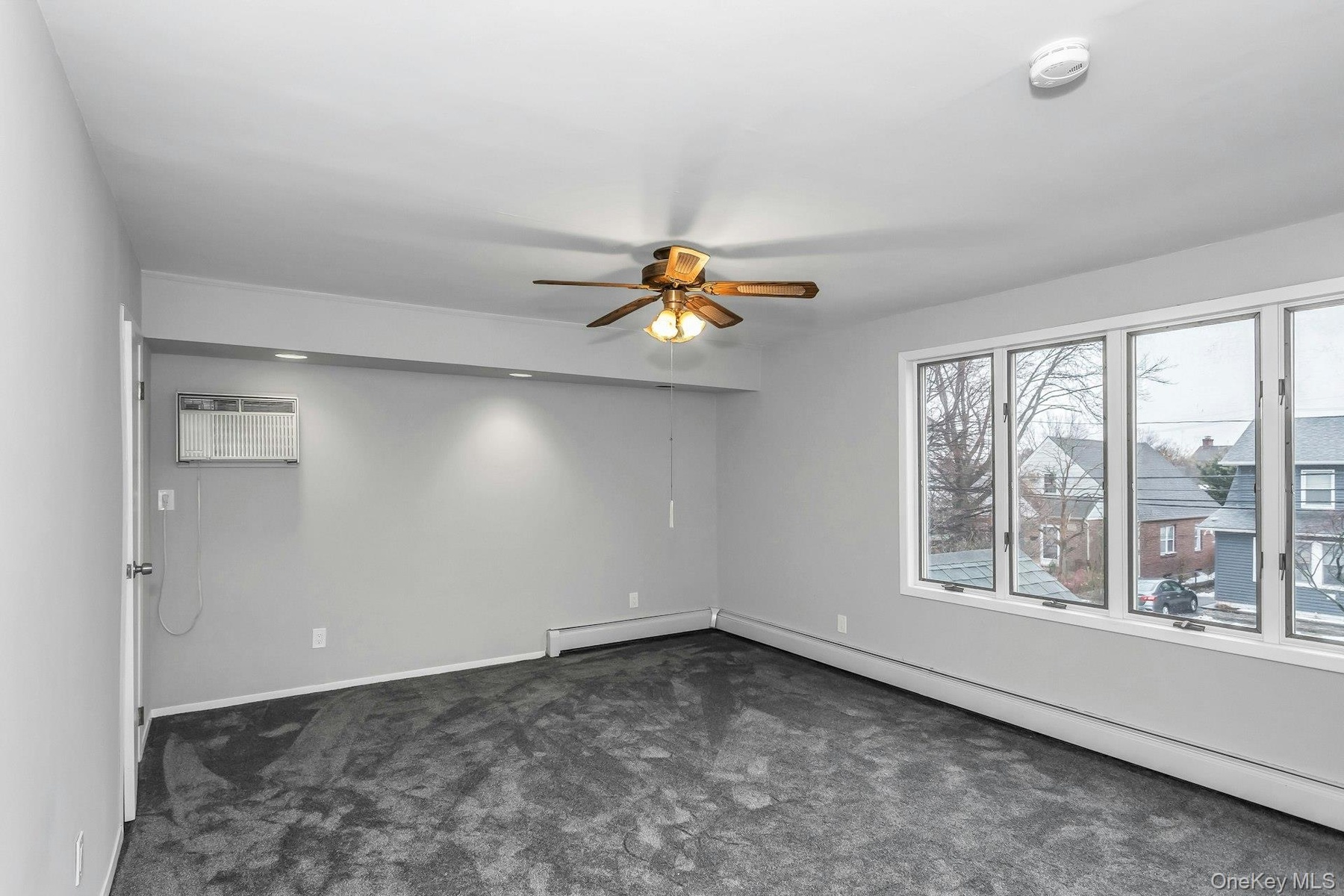 Spacious room featuring light gray walls, dark gray carpet, a multi-pane window, a ceiling fan with integrated lighting, and an air conditioning unit
