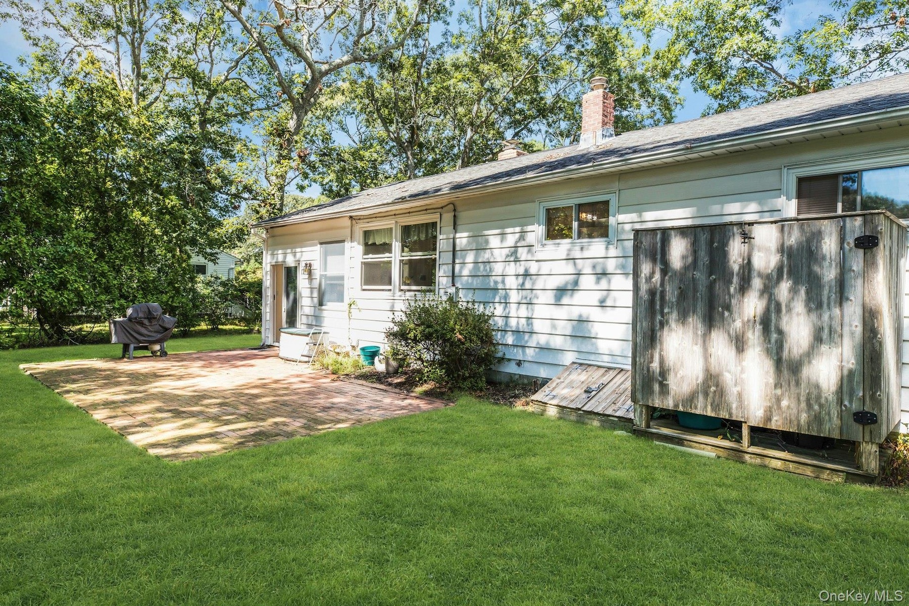 Expansive green lawn, brick paver patio, mature trees, white horizontal siding, and a red brick chimney