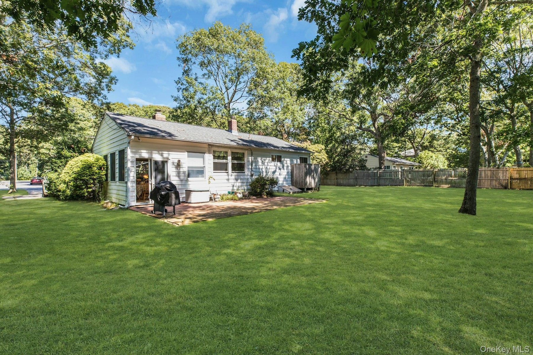 Expansive green lawn, mature trees, white siding exterior, brick patio, and a privacy fence
