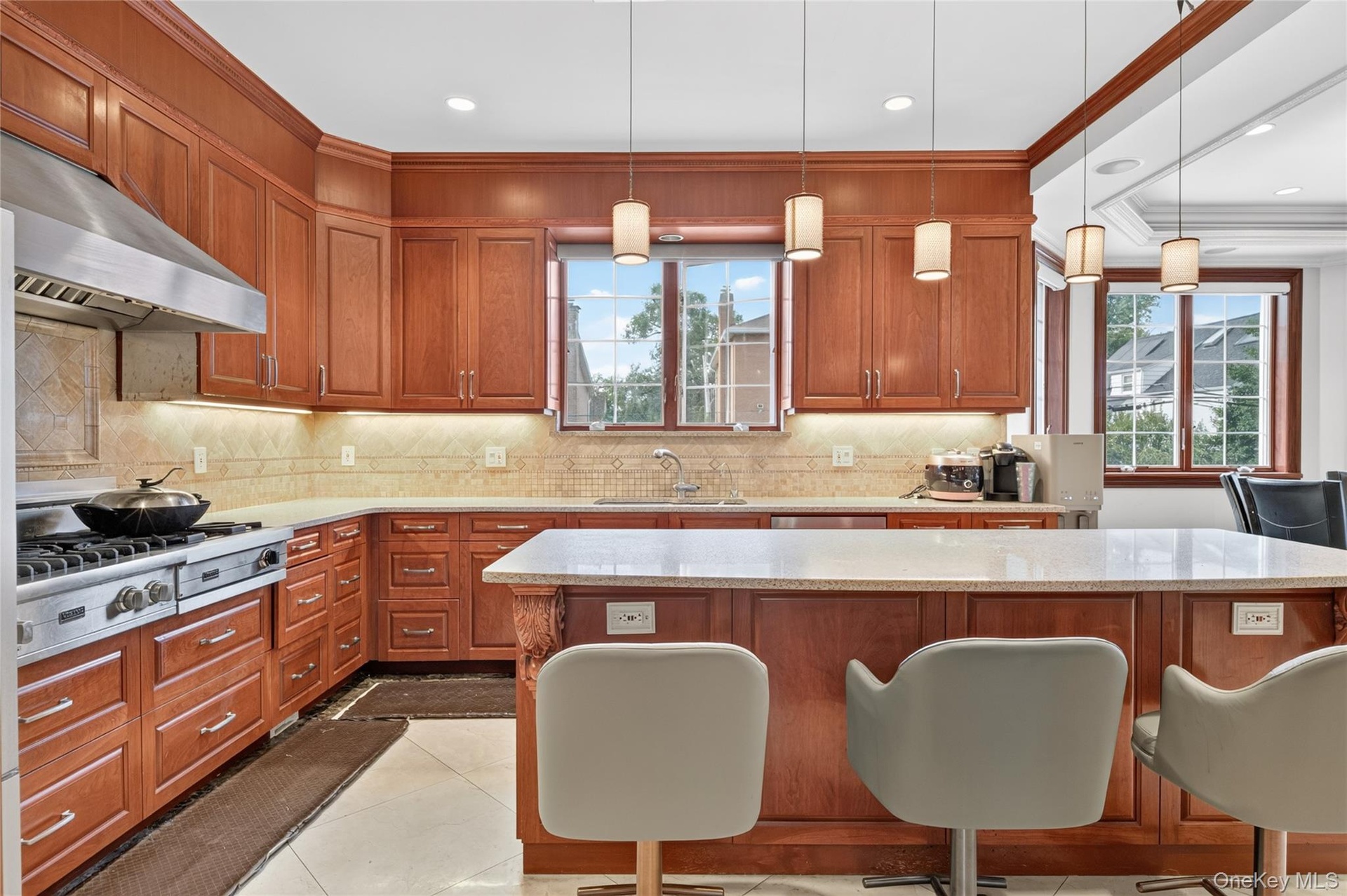 Wood cabinetry, light-toned backsplash, stainless steel range with hood, island with light-toned countertop, and pendant lighting